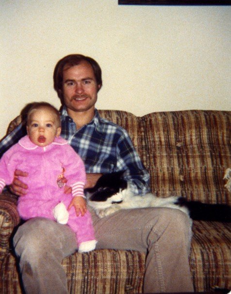 A photo of Emily as a toddler in a onesie, held by her dad on a brown couch. A white and black cat is also in the photo.