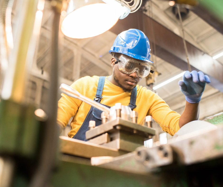 A Black man wearing a hardhat and gloves is working with a piece of manufacturing equipment
